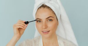 woman in white top and head towel applying mascara on eyelashes