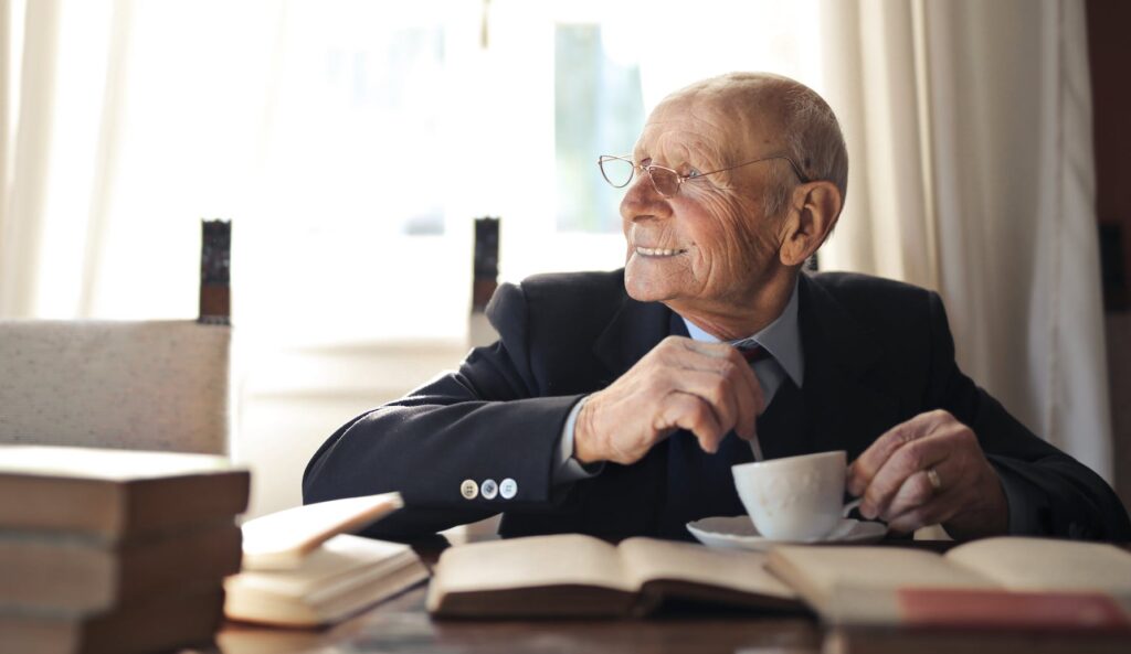 senior man drinking hot beverage while sitting at table with books